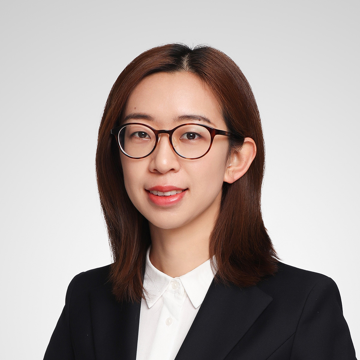 The person in the image is wearing a black blazer over a white collared shirt. They have straight, shoulder-length brown hair. The background is plain and light-colored, likely gray or white. The face is blurred, so no facial features are visible. The overall appearance suggests a professional or formal setting.