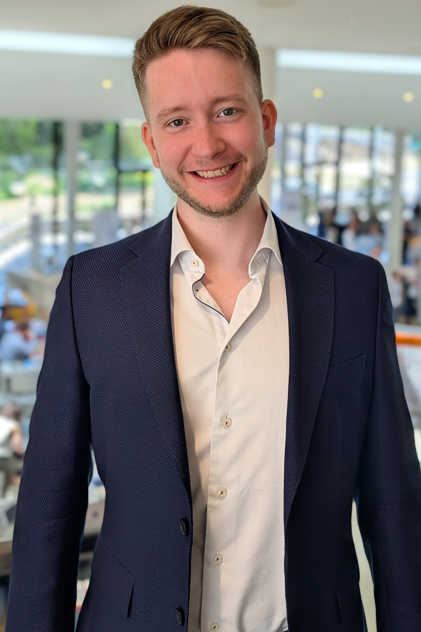 The image shows a person in a navy blazer and light dress shirt standing indoors, likely in a professional or formal setting. The face is obscured by a solid rectangle, so I can’t identify the person.

If you want, I can also help with:
- describing the outfit in more detail
- assessing the photo’s composition or lighting
- suggesting edits for a professional headshot