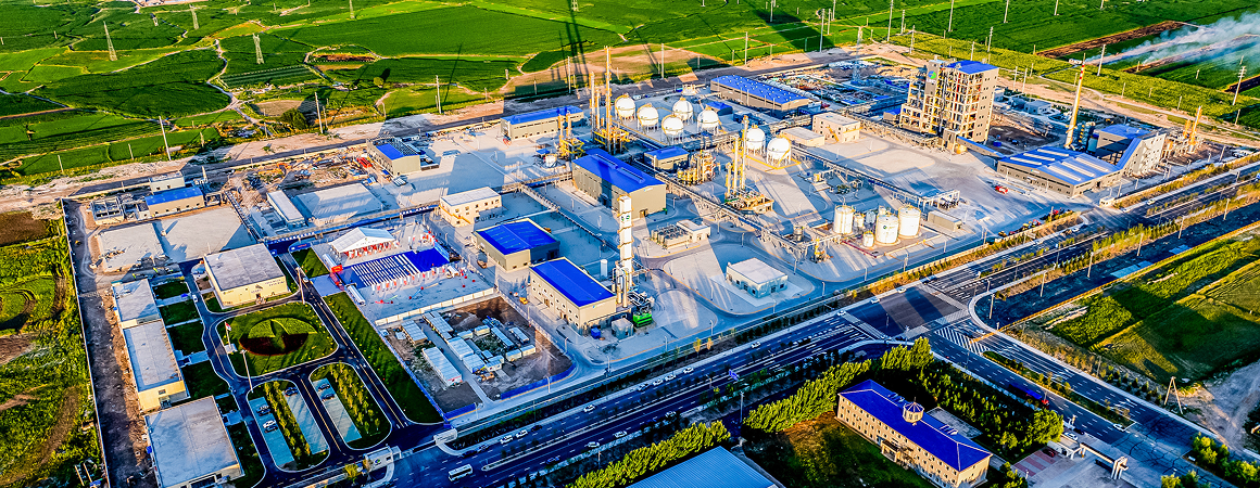 This image shows an aerial view of an industrial complex or factory facility. The facility has multiple buildings, storage tanks, and various industrial equipment. The surrounding area is green with fields, and there are roads and vehicles visible near the complex. The facility appears to be well-organized with clear pathways and infrastructure. The presence of storage tanks and chimneys suggests it might be related to chemical processing, oil refining, or another heavy industry.