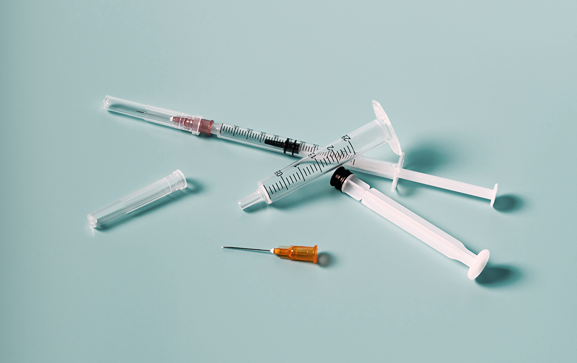 The image shows three syringes and one needle cap on a light blue surface. Two of the syringes are empty, and one contains a small amount of liquid. The needle cap is transparent and is placed near the syringes. The syringes are arranged in a somewhat scattered manner.