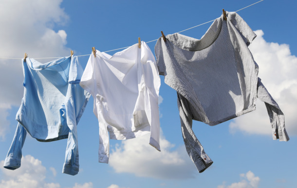 The image shows three shirts hanging on a clothesline against a blue sky with some clouds. The shirts are held by clothespins. The shirts are of different colors: one is light blue, one is white, and one is gray with stripes.