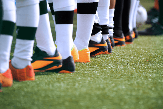 The image shows a row of soccer players standing on a field, with a close-up focus on their lower legs and brightly colored cleats.