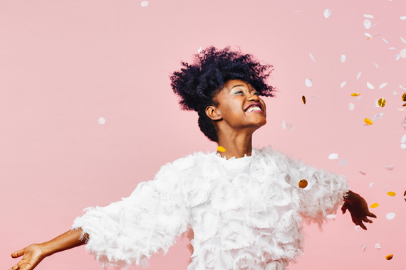 The person in the image is wearing a white textured top with a fluffy or feather-like design. They have short, curly hair and are standing against a pink background. Their arms are extended outward, and there are white and gold confetti pieces floating around them, suggesting a celebratory or joyful moment.