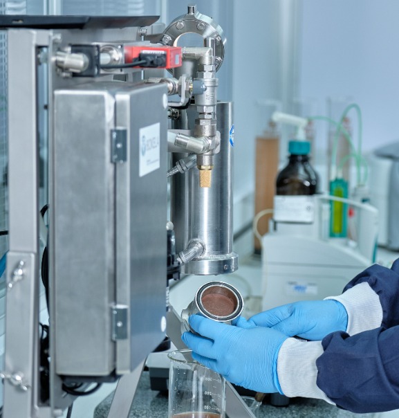 The image shows a person wearing blue gloves handling a metallic component in a laboratory or industrial setting. The component appears to be part of a larger machine or apparatus, possibly related to filtration or fluid processing, given the presence of pipes and a cork stopper. The background includes laboratory equipment and containers, suggesting a controlled environment for scientific or technical work.
