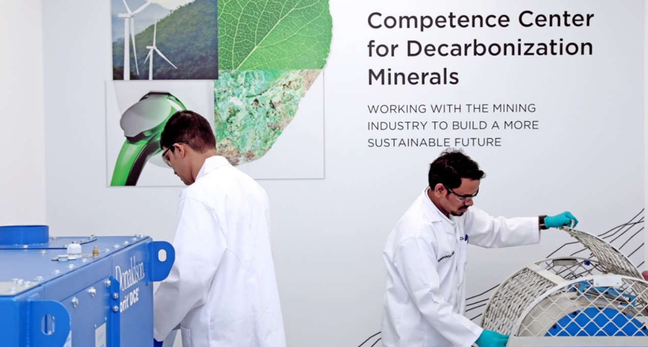 Two scientists in white lab coats working with machinery under a sign that reads "Competence Center for Decarbonization Minerals" promoting sustainable mining.