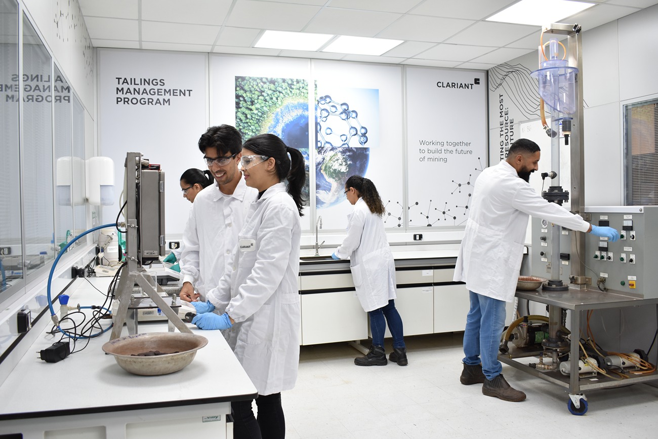 The image shows a laboratory setting where five individuals are engaged in scientific or technical work. They are all wearing white lab coats and blue gloves, indicating adherence to safety and hygiene protocols. The lab is equipped with various instruments and machinery, including a device on the right side that one person is operating. The background features posters with text related to mining, such as "TAILINGS MANAGEMENT PROGRAM" and "Working together to build the future of mining," suggesting that the lab's focus is on mining research or management. The environment is clean and organized, with ample workspace and storage cabinets.