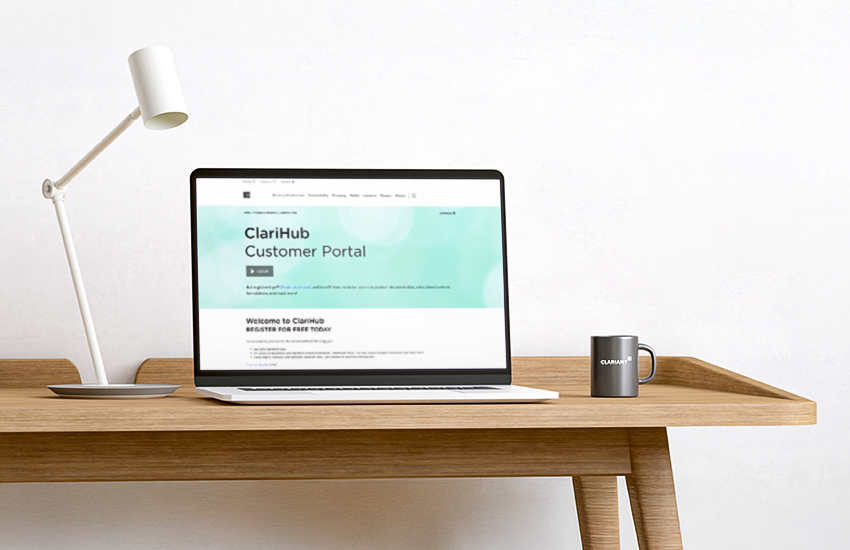The image shows a wooden desk with a modern white desk lamp on the left side, a laptop in the center displaying a webpage titled "ClariHub Customer Portal," and a black coffee mug with the word "CLARIANT" printed on it on the right side. The background is a plain white wall. The overall setting appears to be a clean and minimalistic workspace.