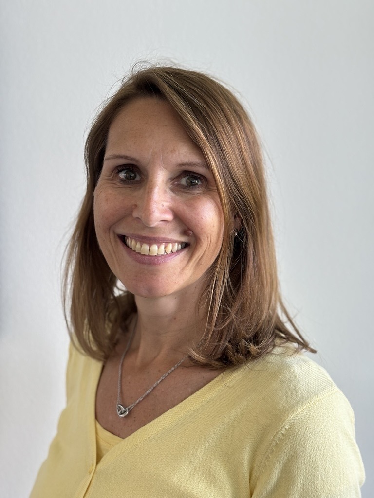 The image shows a person wearing a yellow top and a silver necklace with a heart-shaped pendant. The hair is styled in a medium-length cut with a side part. The background is plain and neutral, which helps to focus attention on the clothing and accessories.