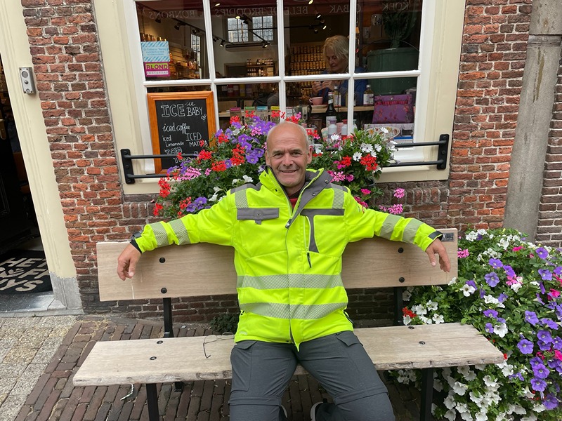 The image shows a person sitting on a bench in front of a brick building with a window. The individual is wearing a bright yellow high-visibility jacket with reflective stripes and dark pants. Behind the person, there is a display of colorful flowers, and a chalkboard sign advertising iced coffee and iced matcha. The setting appears to be a quaint, possibly European street with a cozy atmosphere.