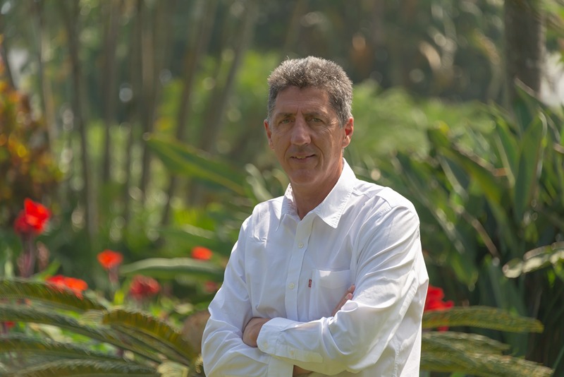 The person is wearing a white button-up shirt and is standing in a lush, green outdoor setting with vibrant red flowers and various plants in the background. The lighting suggests it is a sunny day.
