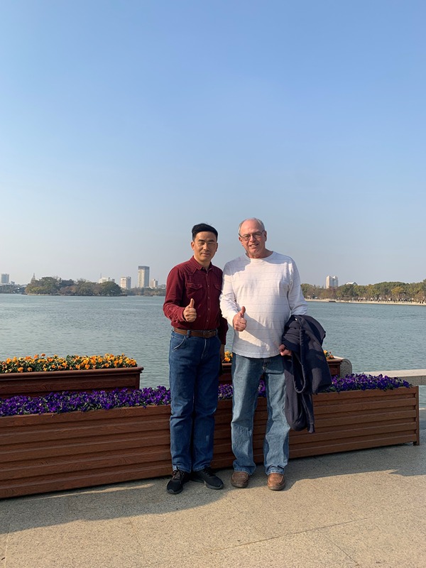 The image shows two individuals standing side by side in front of a body of water. They are giving a thumbs-up gesture. The person on the left is wearing a maroon shirt and blue jeans, while the person on the right is wearing a white sweater and blue jeans, holding a jacket. There are flower boxes with colorful flowers in the foreground, and buildings and trees can be seen in the background across the water. The sky is clear and blue, suggesting a pleasant day.