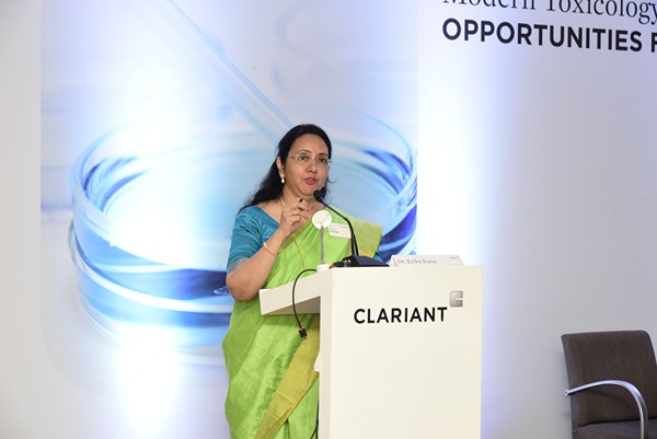 The image shows a person standing at a podium labeled "CLARIANT," delivering a speech or presentation. The backdrop features a scientific theme, possibly related to modern toxicology, as indicated by the partial text visible. The individual is wearing a green and blue saree, suggesting a formal or professional event.