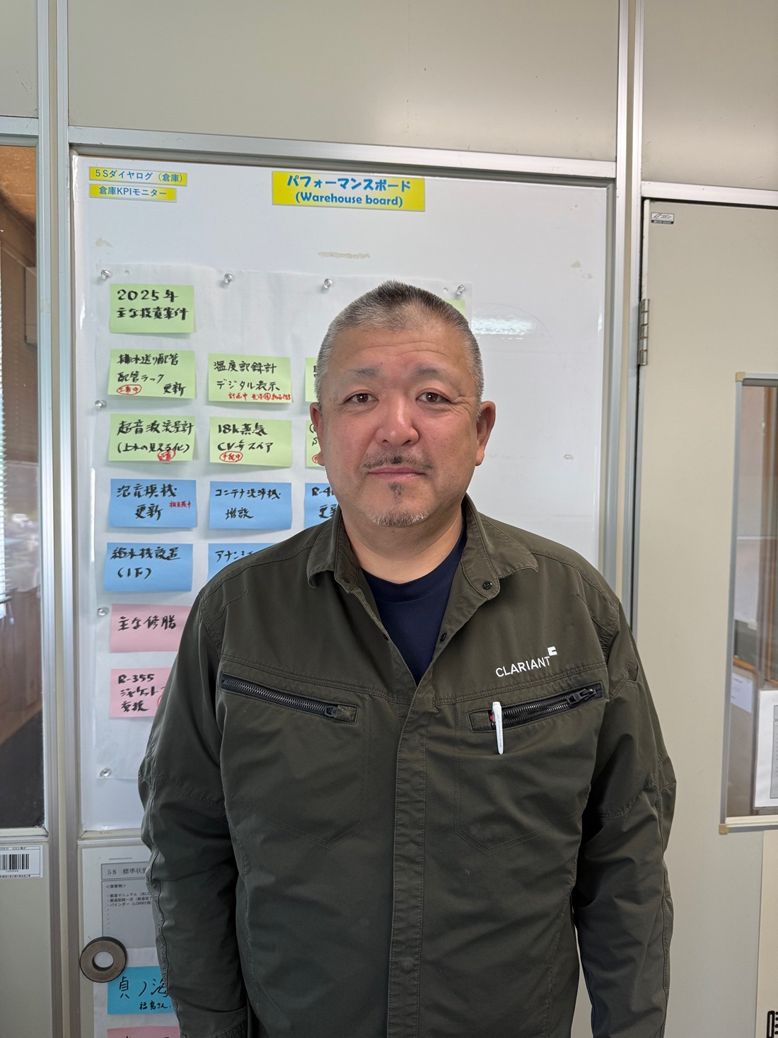 The image shows a person standing in front of a performance board labeled "Warehouse board." The board contains various notes and plans written in Japanese, indicating goals and strategies for the year 2025. The person is wearing a jacket with the logo "Clariant," suggesting they might be associated with the company Clariant, which is known for specialty chemicals. The setting appears to be an office or a workplace environment.