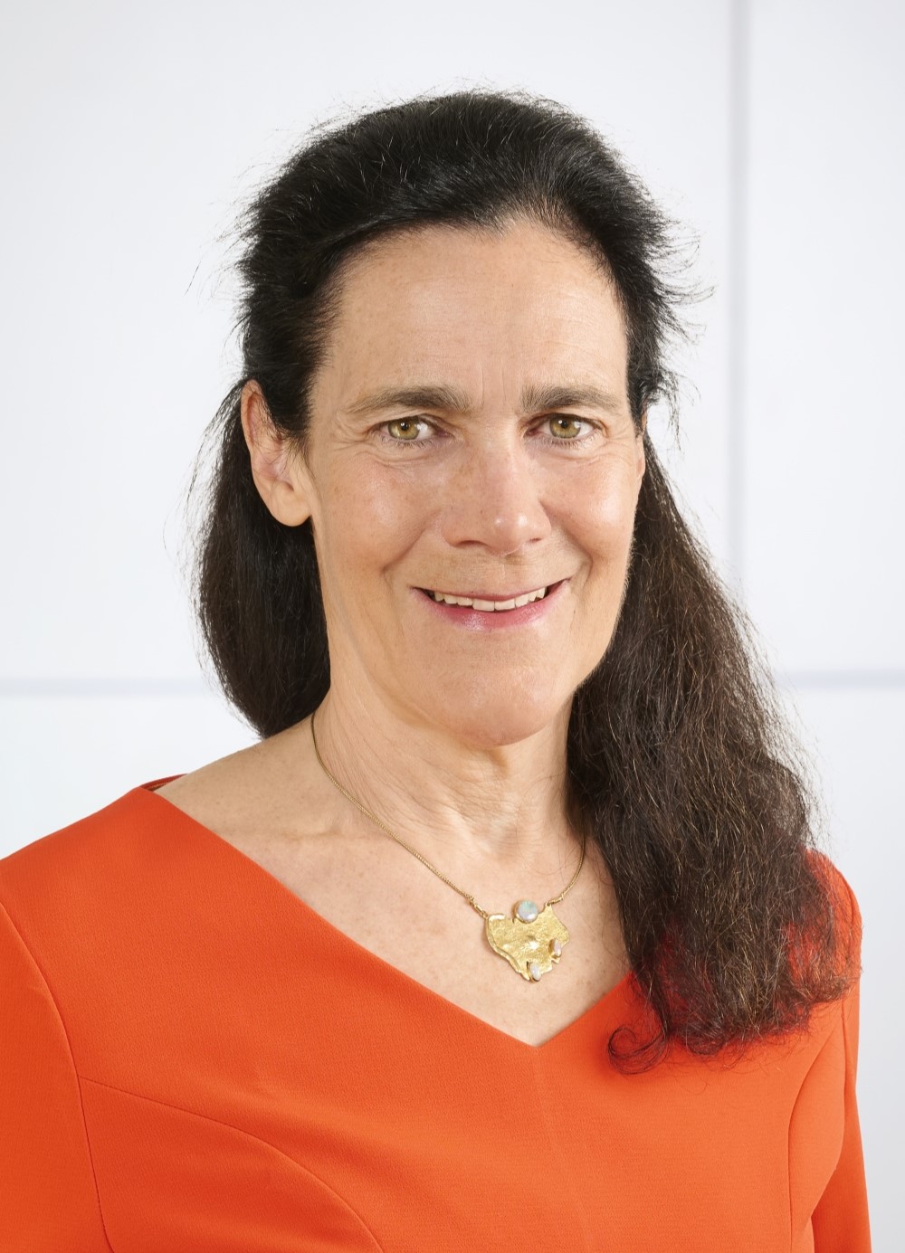 Person with long dark hair wearing an orange top and a gold necklace with a pendant, photographed against a plain light background.