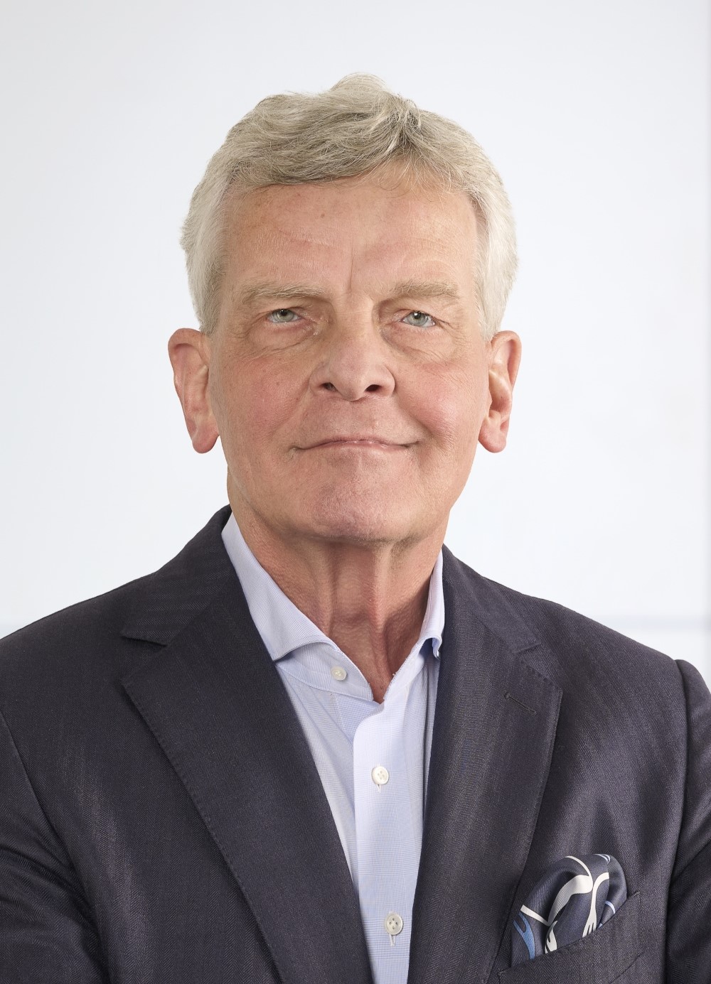 Man with gray hair wearing a dark suit jacket, light blue shirt, and patterned pocket square against a plain white background.