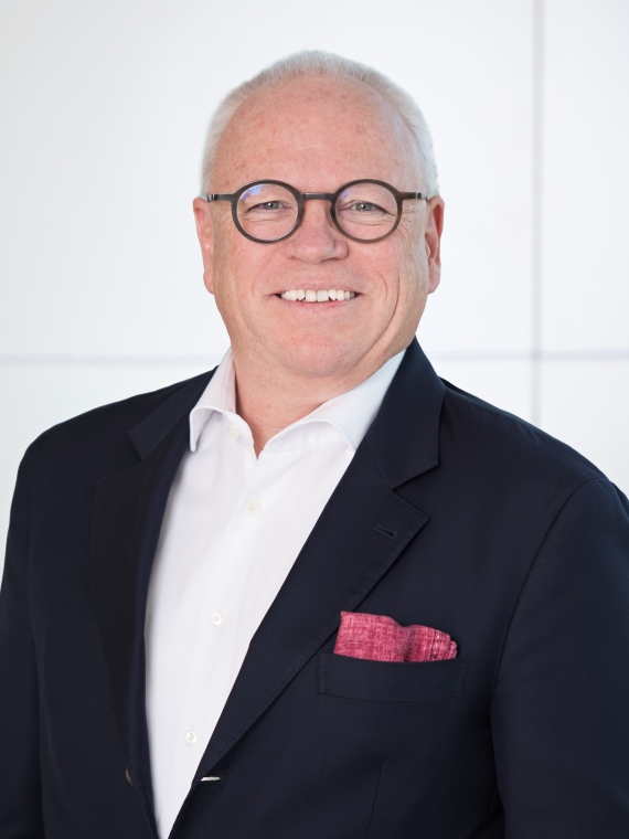 The person in the image is wearing a dark navy blazer over a white dress shirt. There is a red pocket square neatly placed in the breast pocket of the blazer. The background appears to be a plain, light-colored wall with subtle horizontal lines. The overall attire suggests a formal or business setting.