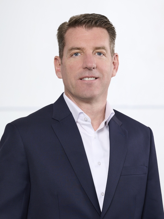 The person in the image is wearing a dark navy blue suit jacket over a white dress shirt. The shirt is buttoned up, but without a tie. The background is plain and light-colored, giving a professional and clean appearance. The hairstyle is neatly combed back.