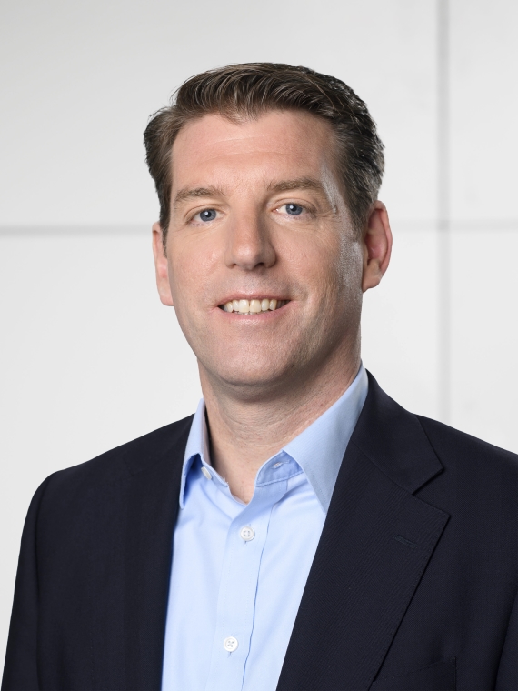 The individual is wearing a dark suit jacket over a light blue dress shirt. The shirt is buttoned up, and the collar is neatly arranged. The hair is styled in a neat, professional manner. The background is a plain, light-colored wall, suggesting a formal or professional setting.