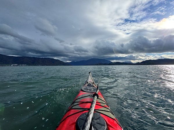 This image shows the front of a red kayak on a body of water, likely a lake or calm sea. The kayak is pointed towards distant mountains under a dramatic sky with a mix of dark clouds and patches of blue. The water appears slightly choppy with small waves. The overall scene suggests an outdoor adventure or kayaking trip in a scenic, natural environment.