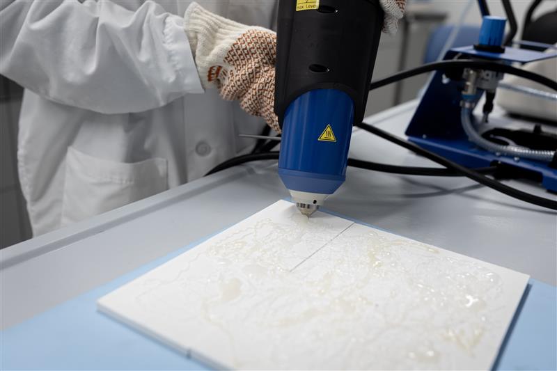 The image shows a person wearing protective gloves and a lab coat operating a laser engraving or cutting machine. The machine is being used on a white tile or similar flat material placed on a work surface. The laser head is positioned close to the tile, and the person appears to be guiding or adjusting the machine. The environment looks like a laboratory or workshop setting.