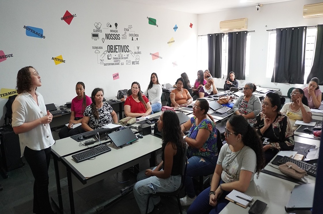 This image shows a classroom or training room setting with a group of people, mostly women, seated and attentively listening to a woman standing at the front, who appears to be the instructor or speaker. The room has several desks with computers and notebooks, indicating a learning or workshop environment.

On the wall behind the instructor, there are motivational words and phrases in Portuguese, such as "NOSSOS OBJETIVOS" (Our Objectives), "CLIENTES" (Clients), "INOVAÇÃO" (Innovation), "ÉTICA" (Ethics), "EXCELÊNCIA" (Excellence), "TECNOLOGIA" (Technology), "VALORES" (Values), "SUSTENTABILIDADE" (Sustainability), and "METAS" (Goals). Additionally, colorful paper cutouts with words like "SUPERACAO" (Overcoming), "ATITUDE" (Attitude), "FOCO" (Focus), and "AGILIDADE" (Agility) are displayed on the walls.

The atmosphere suggests a professional or educational session focused on development, motivation, or skills training.