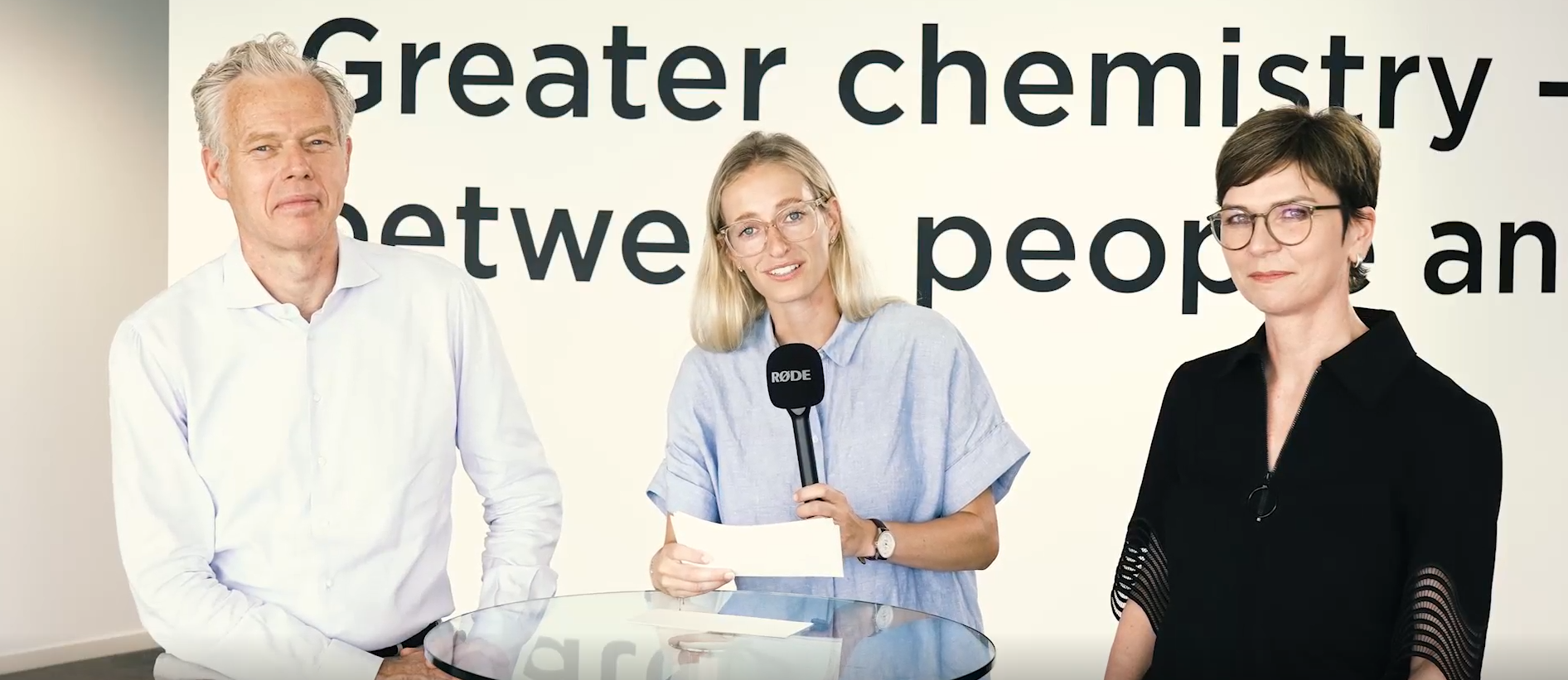 Three people standing behind a glass table, one holding a microphone and a paper, with a wall text in the background.