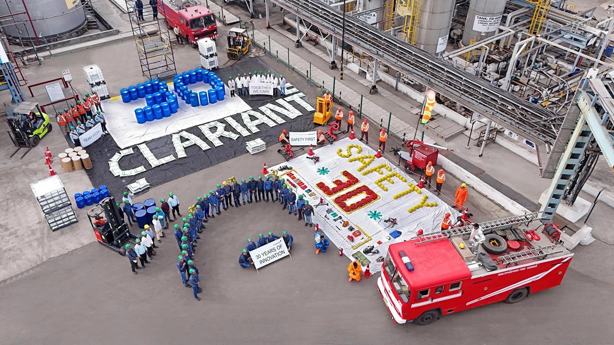 The image shows a group of workers in an industrial setting celebrating a milestone related to safety and innovation. The workers are arranged in a spiral formation, holding a sign that reads "30 YEARS OF INNOVATION." Nearby, there are large displays made from various materials:

- The number "30" is formed using blue barrels.
- The word "CLARIANT" is spelled out using white bags or sacks.
- Another display reads "SAFETY 30" with the word "SAFETY" in yellow and "30" in red, surrounded by safety equipment and tools.
- Additional signs include "TOGETHER WE CARE," "TEAM WORK," and "SAFETY FIRST."

There is a red fire truck parked nearby, and the workers are wearing safety gear such as helmets and vests, indicating a focus on workplace safety. The setting appears to be an industrial plant or factory with tanks and pipelines in the background.