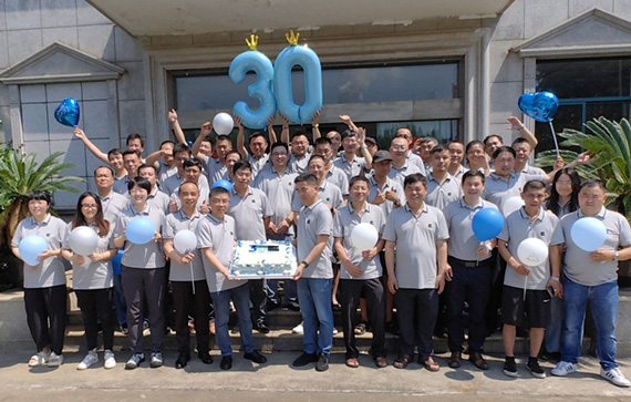 This image shows a large group of people gathered outside a building, celebrating an event. Most of them are wearing matching light gray polo shirts, suggesting they are part of the same organization or team. They are holding balloons in blue and white colors, and there are two large blue balloons shaped as the number "30" at the center, indicating a 30th anniversary or milestone celebration. Some people in the front row are holding a cake, further emphasizing the celebratory nature of the gathering. The atmosphere appears joyful and festive.