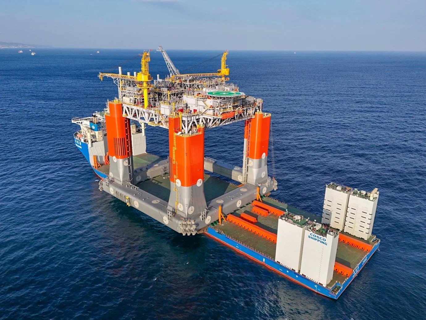 The image shows a large offshore platform being transported on a heavy-lift vessel. The platform has four large cylindrical legs painted orange and gray, supporting a deck with various equipment and a helipad. The vessel carrying the platform is marked with "COSCO SHIPPING," indicating it belongs to the COSCO Shipping fleet, a major Chinese shipping company. The platform is likely used for oil or gas extraction or other offshore industrial purposes. The surrounding environment is open sea with a clear sky.
