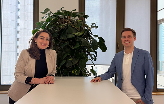 The image shows two people sitting at a white table in an office-like setting. There is a large green plant in the background between them. The person on the left is wearing a beige blazer over a black top, and the person on the right is wearing a blue blazer over a white shirt. Both individuals are facing the camera, and there are large windows behind them letting in natural light.