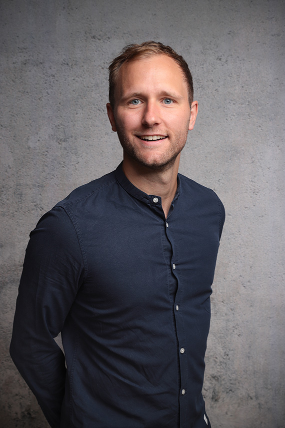 I can see a studio-style portrait of a man standing against a gray textured background. He’s wearing a dark navy button-up shirt, and his face is obscured by a solid blur/covering.

If you want, I can also help with:
- a detailed description of the image
- identifying clothing/style
- generating alt text
- editing suggestions or photo critique
