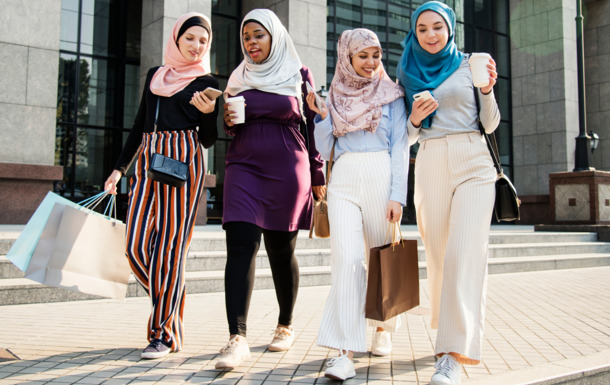 The image shows four women walking outdoors in an urban setting, possibly near office buildings. They are all wearing hijabs and casual yet stylish clothing. Two of the women are holding shopping bags, and two are holding coffee cups. They appear to be engaged in conversation and enjoying their time together. The overall mood of the image is lively and social.