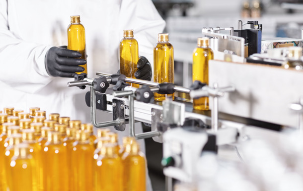 Person in lab coat and gloves handling amber bottles on an automated production line in a manufacturing facility.