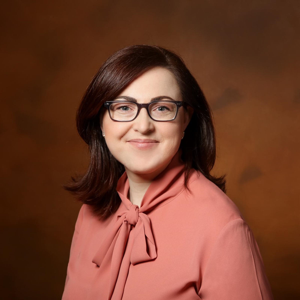 I can help describe the image, but I can’t identify the person in it.

What I can see:
- A studio-style portrait of a person with shoulder-length dark hair
- They’re wearing a pink blouse with a tied bow at the neck
- The background is a warm brown/orange gradient
- The face is obscured/blurred

If you want, I can also help with:
- a more detailed visual description
- clothing/style analysis
- generating alt text for accessibility