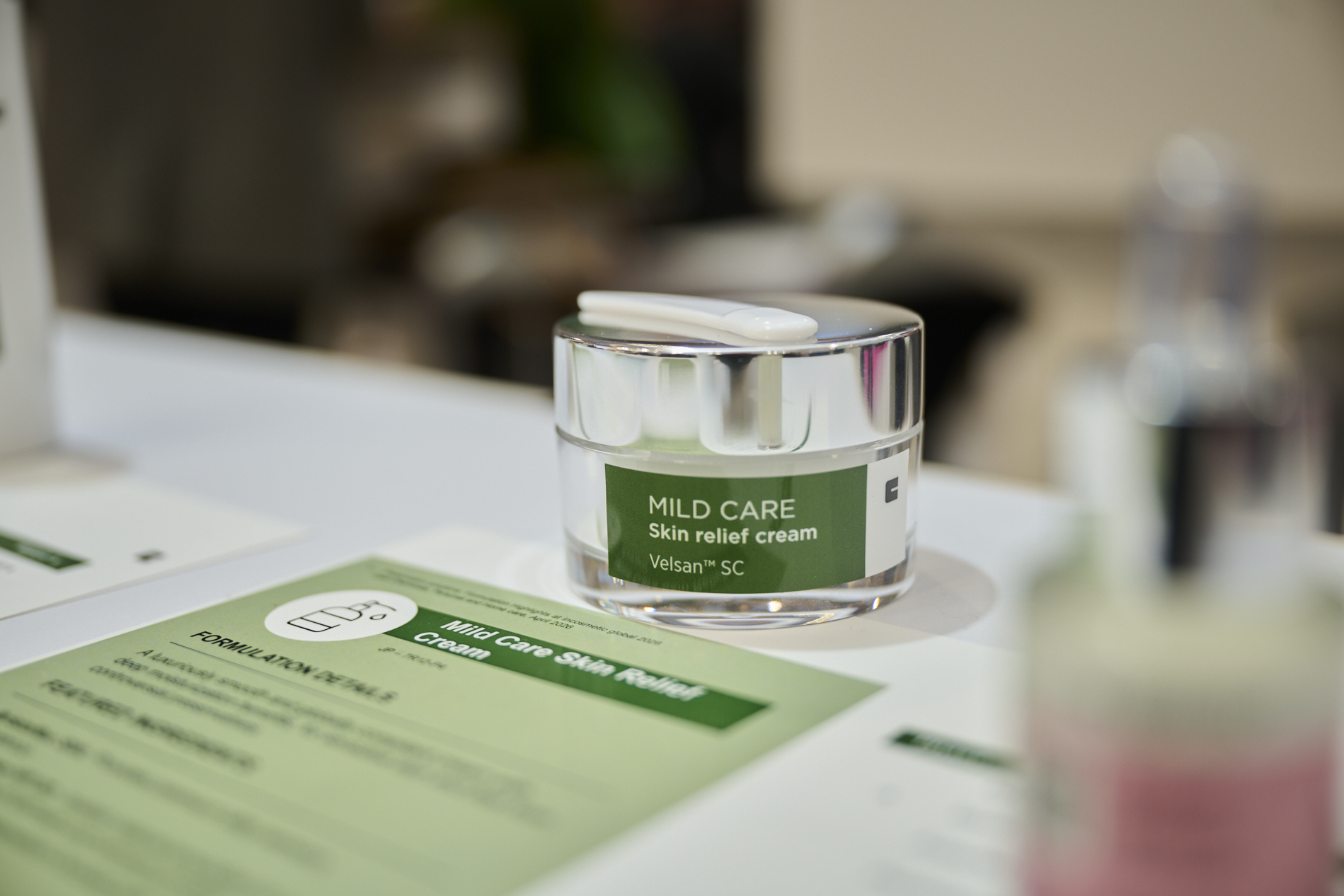 The image shows a small cosmetic jar labeled **“MILD CARE Skin relief cream”** with **“Velsan™ SC”** on the front. It’s placed on a table next to a printed product sheet or brochure, likely in a product presentation or lab/marketing setting. The background is blurred, with another out-of-focus bottle visible on the right.